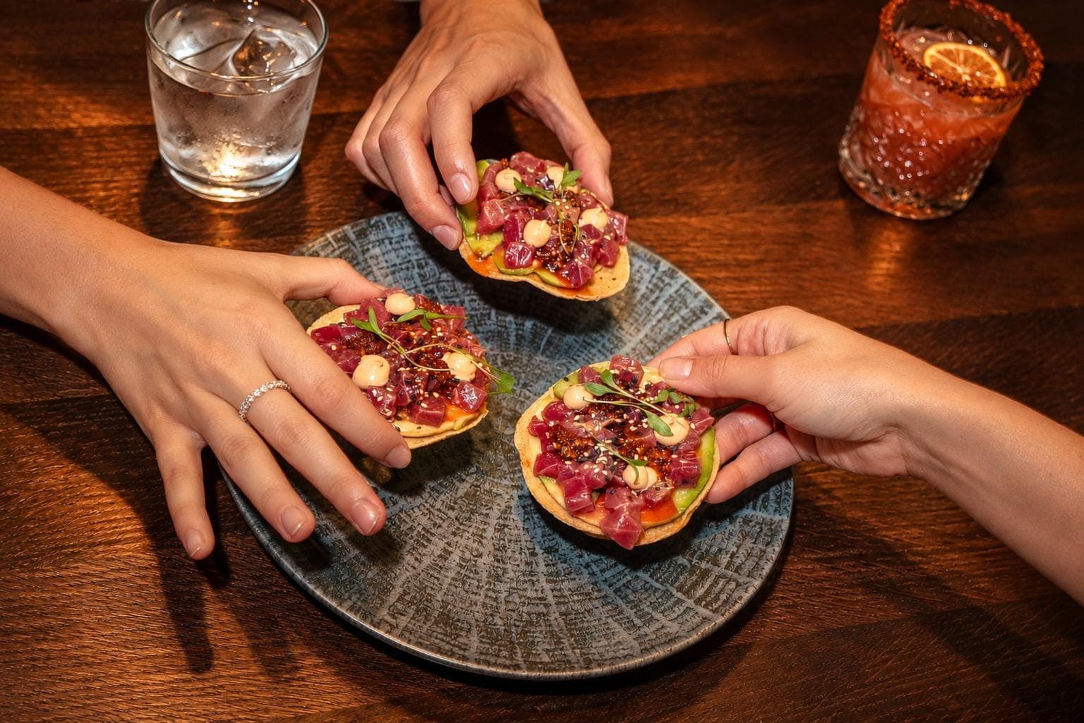 Tres manos se llevan las tostadas de atún, con un cóctel con borde de chile en la mesa